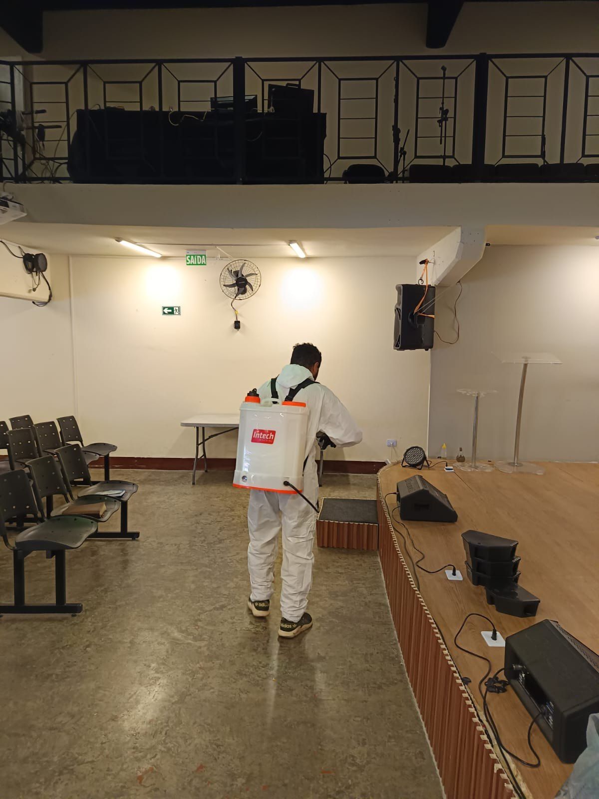 Técnico com pulverizador costal aplicando produto em salão com cadeiras e palco